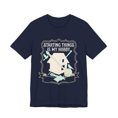 Starting Things Is My Hobby tee flat front, navy unisex tee, a half-built birdhouse with brush and yarn