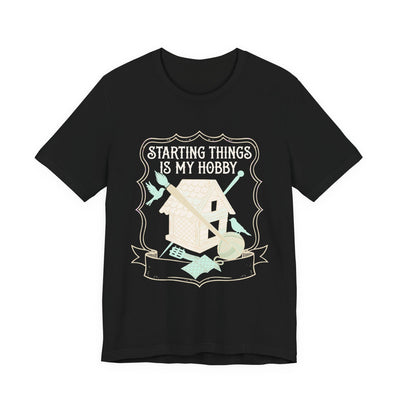 Starting Things Is My Hobby tee flat front, black unisex tee, a half-built birdhouse with brush and yarn