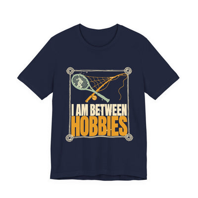 I Am Between Hobbies tee flat front, navy unisex tee, a dusty tennis racket and fishing rod crossed