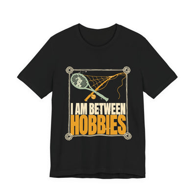 I Am Between Hobbies tee flat front, black unisex tee, a dusty tennis racket and fishing rod crossed