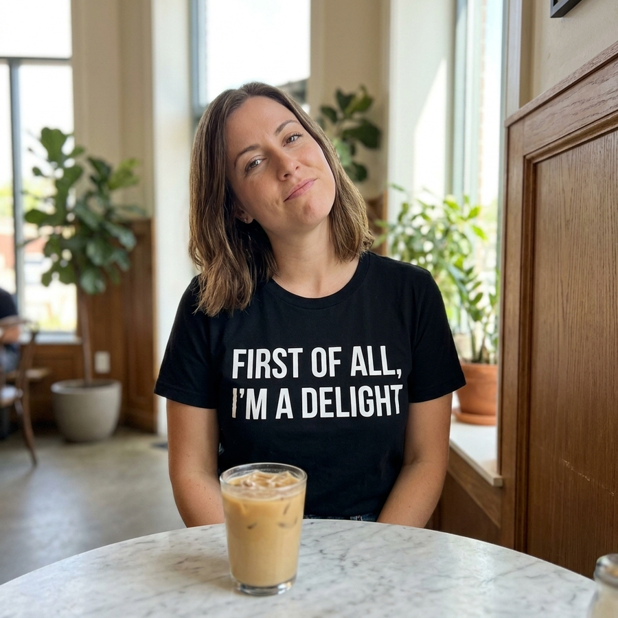Confident woman in bright cafe wearing black 'First of All I'm a Delight' statement t-shirt with bold white text